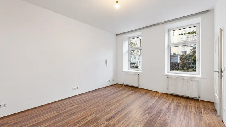 Geräumiges Wohnzimmer mit Holzboden und zwei großen Fenstern, die viel Tageslicht hereinlassen.