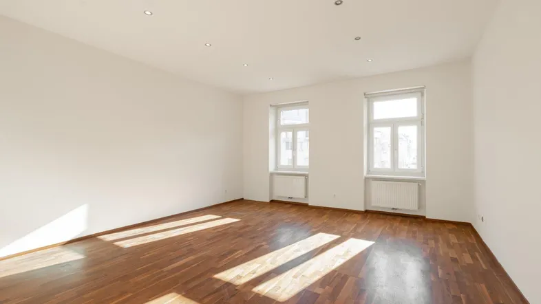 Heller, leerer Raum mit Holzboden und zwei Fenstern, durch die Sonnenlicht fällt.