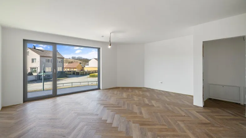Helles, unmöbliertes Wohnzimmer mit Fischgrätparkett und großem Fenster mit Blick auf die Umgebung.