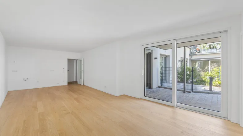 Helles, unmöbliertes Wohnzimmer mit Holzboden und Zugang zu einer gepflasterten Terrasse.
