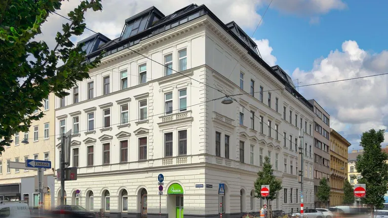 Historisches Eckgebäude mit weißer, reich verzierter Fassade und modernem Mansarddach in urbaner Umgebung.