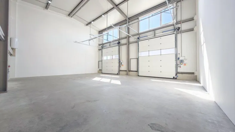 Geräumige, leere Lagerhalle mit hohen Decken, Betonboden und großen Rolltoren, die Tageslicht hereinlassen.