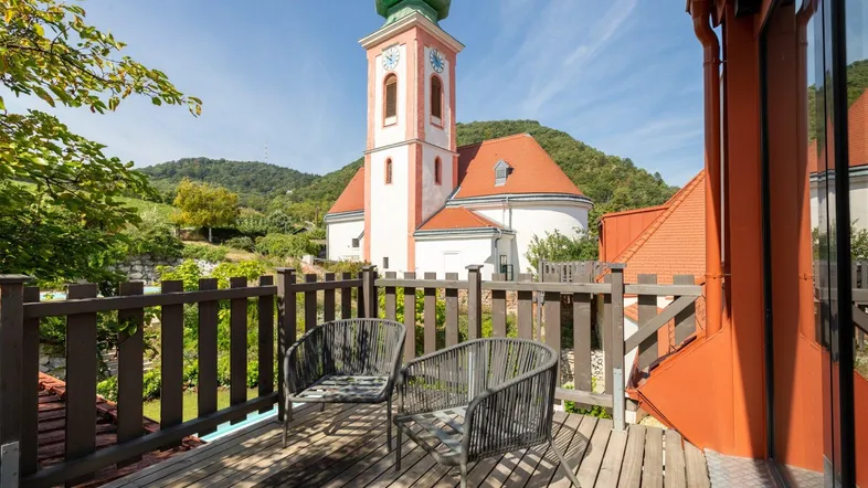 Gemütlicher Balkon mit Holzboden und Blick auf die Kirche sowie die umliegenden Weinberge.