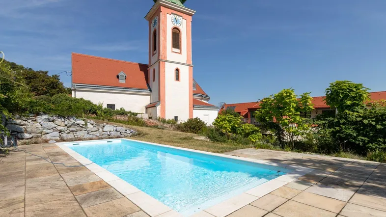 Außenpool mit gefliester Terrasse und historischer Kirche im Hintergrund unter blauem Himmel.