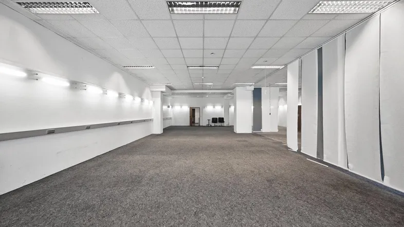 Großer, leerer Gewerberaum mit Teppichboden, abgehängter Decke und Wandbeleuchtung, bereit zur individuellen Gestaltung.