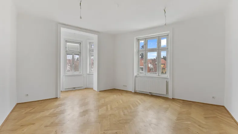 Helles Zimmer mit Fischgrätparkett und großen Fenstern, das viel Tageslicht hereinlässt.