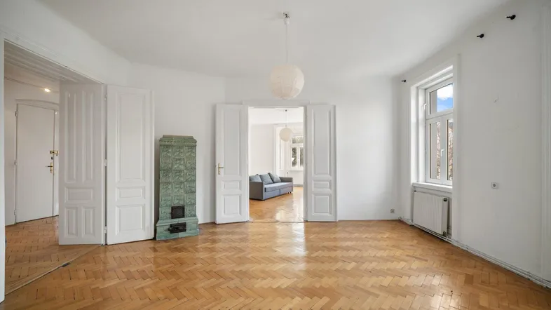 Geräumiges Wohnzimmer mit Fischgrätparkett und einem traditionellen Kachelofen, der zu einem weiteren Raum führt.