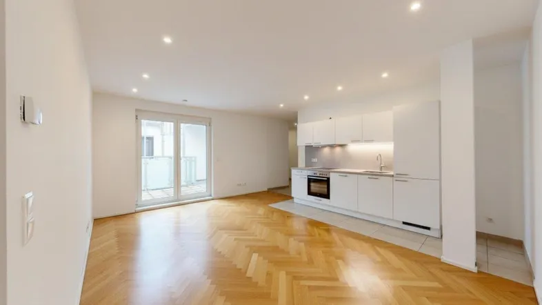 Heller offener Wohnbereich mit Fischgrätparkett, integrierter Küche und Zugang zu einem Balkon.