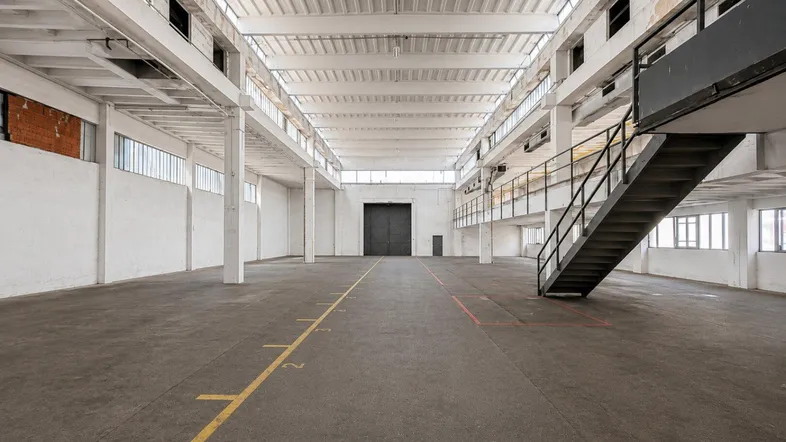 Geräumige Industriehalle mit hohen Decken, großen Fenstern und einer internen Treppe zu einer Empore.