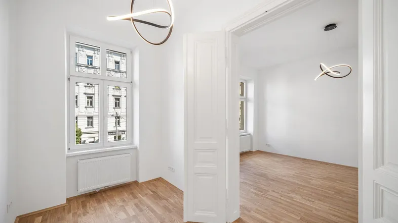 Helles Zimmer mit Parkettboden und Blick auf die Straße, verbunden mit einem weiteren Raum.