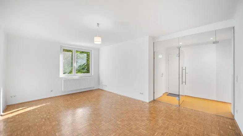 Helles, leeres Zimmer mit Parkettboden und großen Fenstern, durch eine Glaswand abgetrennt.
