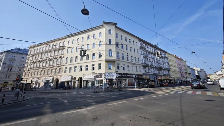 Blick auf ein Eckgebäude an einer belebten Straße mit Geschäften im Erdgeschoss und Wohnungen darüber.