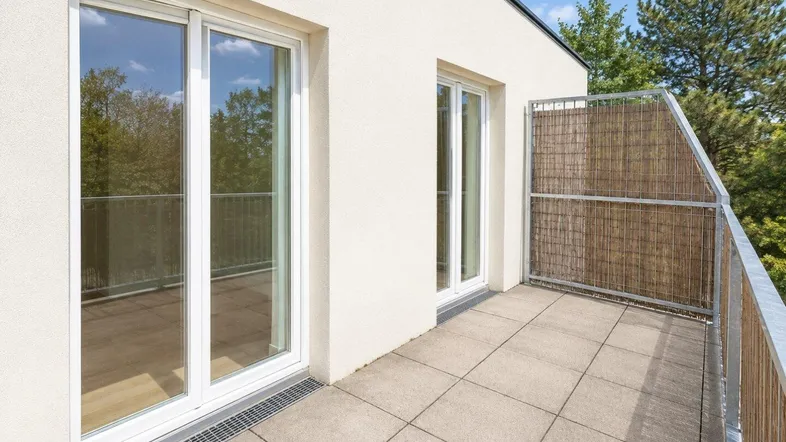 Heller Balkon mit Fliesenboden, Schiebetüren und Sichtschutz, umgeben von viel Grün.