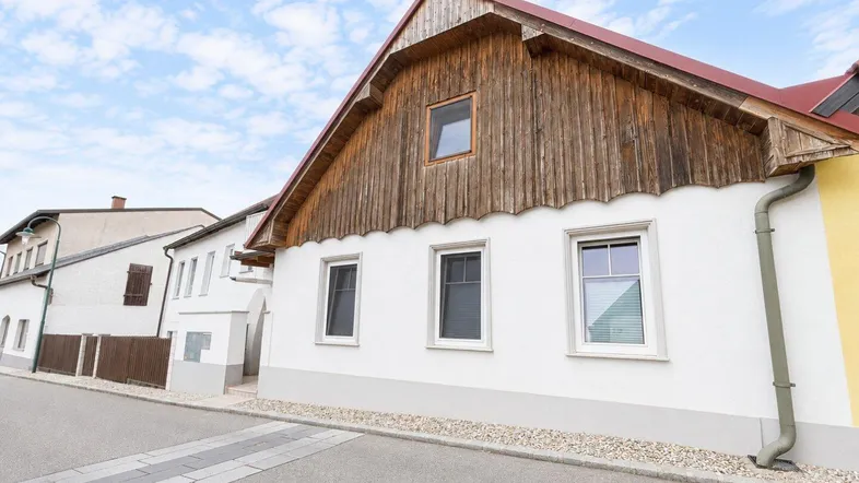 Traditionelles Haus mit weißer Fassade und Holzverkleidung unter einem roten Satteldach an der Straße.