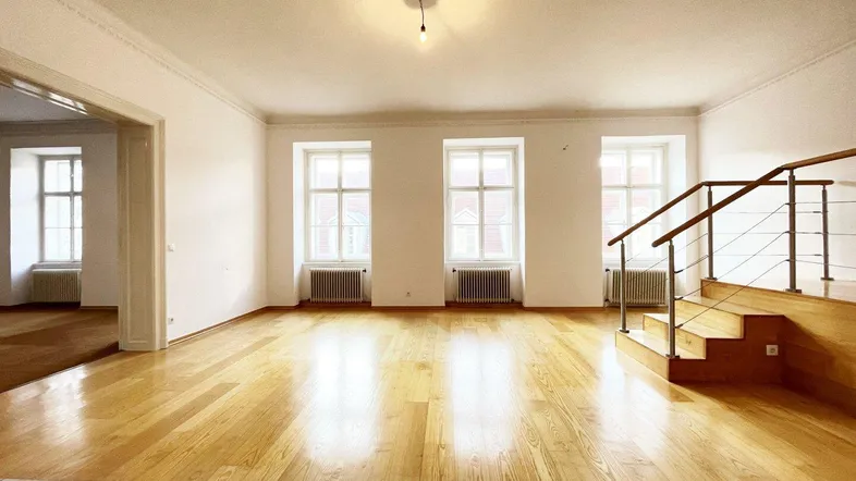 Helles Wohnzimmer mit Holzboden, hohen Fenstern und einer Treppe, die zu einem weiteren Bereich führt.