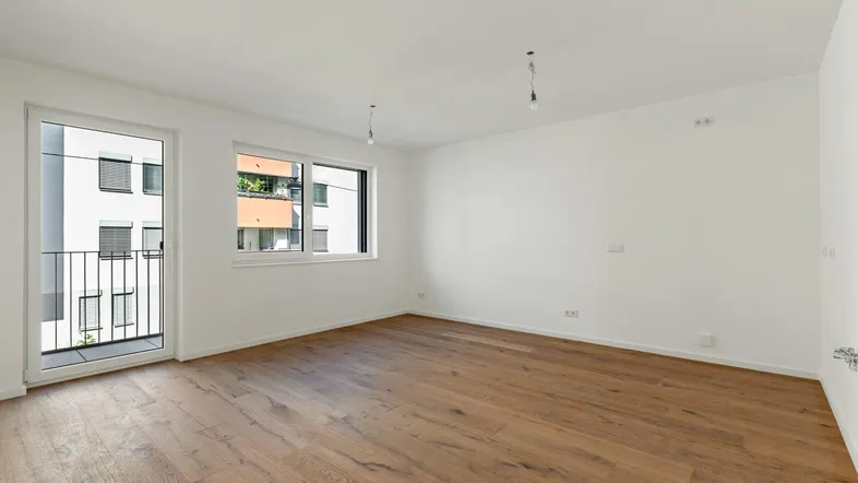Helles, leeres Zimmer mit Holzboden und Zugang zu einem kleinen Balkon.