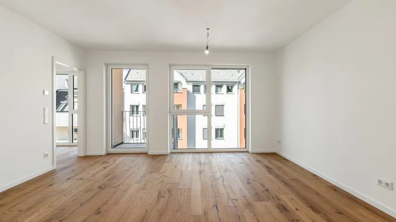 Helles, leeres Wohnzimmer mit großen Fenstern, Holzboden und Zugang zu einem französischen Balkon.