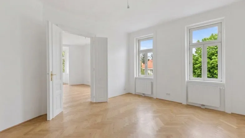 Heller, leerer Raum mit Fischgrätparkett und hohen Fenstern, der viel Tageslicht hereinlässt.