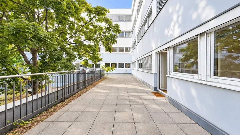 Große, geflieste Terrasse mit Metallgeländer und Blick auf grüne Bäume und das Gebäude.