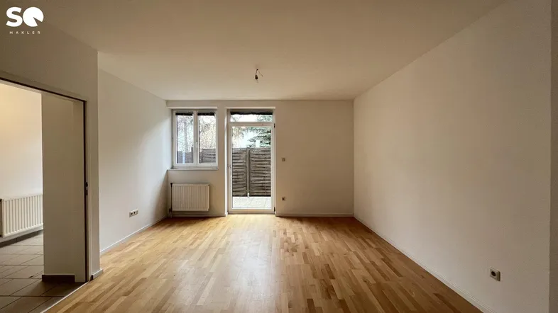 Helles, leeres Wohnzimmer mit Parkettboden und Zugang zu einem kleinen Außenbereich.