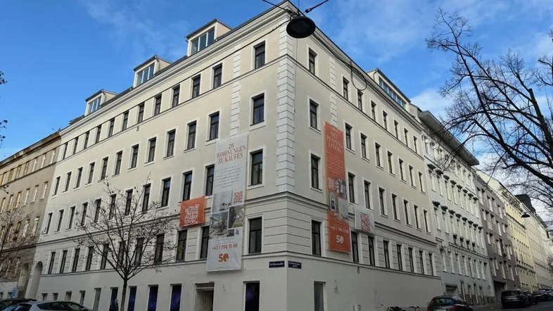 Mehrstöckiges Eckgebäude mit heller Fassade, vielen Fenstern und Mansarddach in städtischer Umgebung.