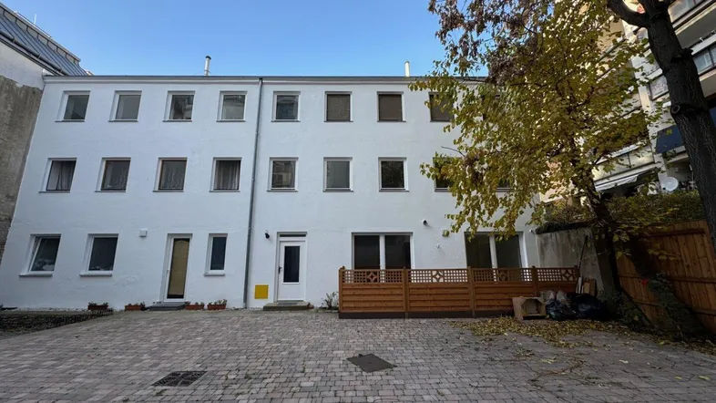 Weißes Mehrfamilienhaus mit gepflastertem Innenhof und kleiner Holzterrasse im Erdgeschoss.