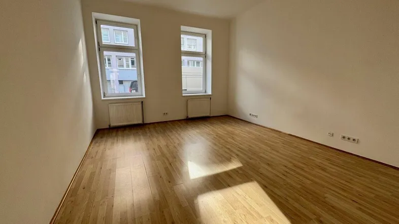 Helles, leeres Zimmer mit Holzfußboden und zwei Fenstern, bereit zur individuellen Gestaltung.