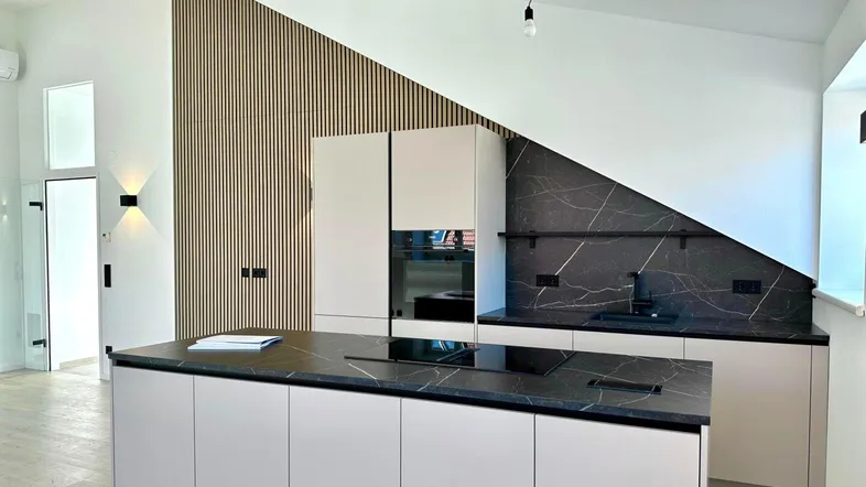 Moderne Küche mit Kücheninsel, dunkler Marmorarbeitsplatte und Holzlamellenwand unter Dachschräge.