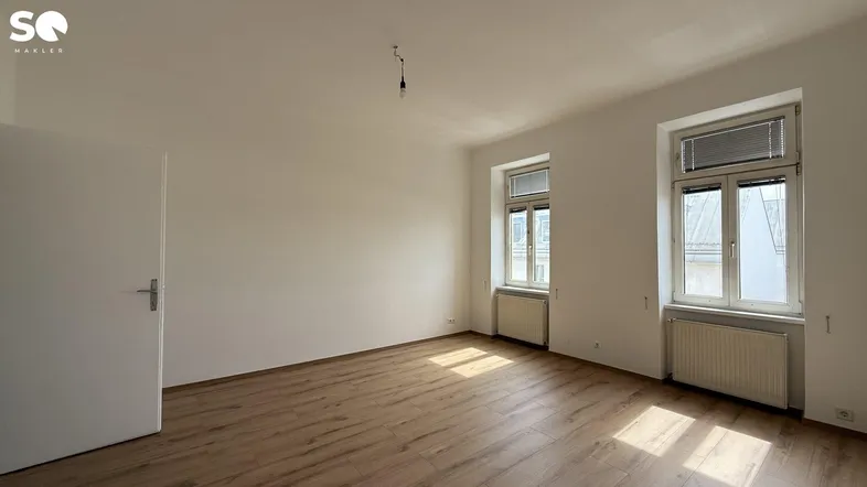 Heller Raum mit Holzboden und zwei Fenstern, eine weiße Tür führt in den Flur.