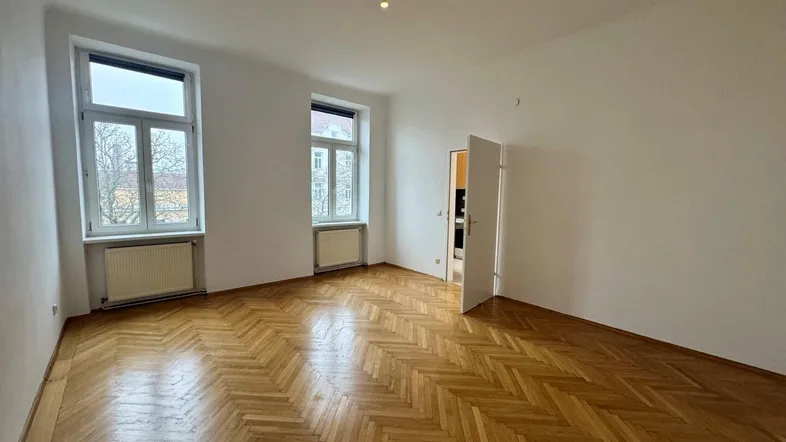 Helles Zimmer mit Fischgrätparkettboden und großen Fenstern, ideal als Wohn- oder Schlafzimmer.