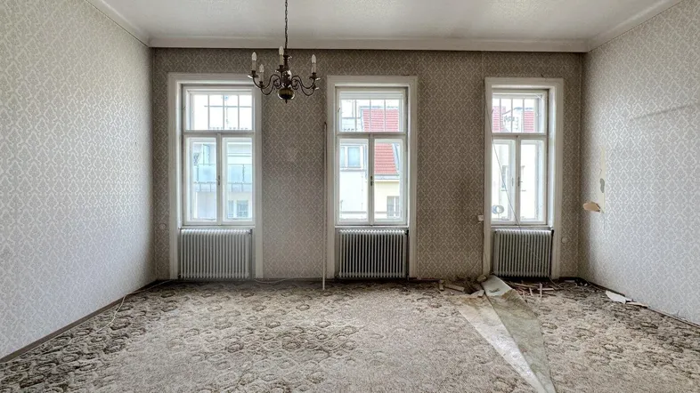 Geräumiges Zimmer mit drei hohen Fenstern, Kronleuchter und Renovierungsbedarf.