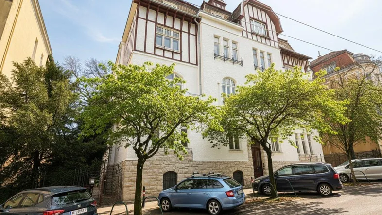 Traditionelles Gebäude mit weißer Fassade, Fachwerkdetails und parkenden Autos auf der Straße.