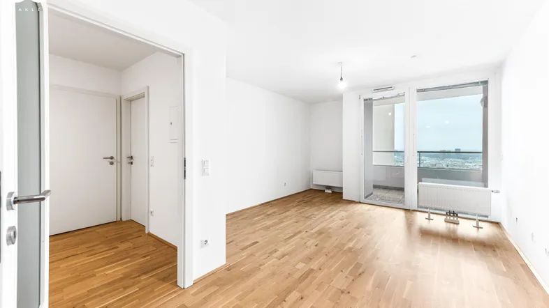 Helles, leeres Wohnzimmer mit Parkettboden und Zugang zu einem Balkon.