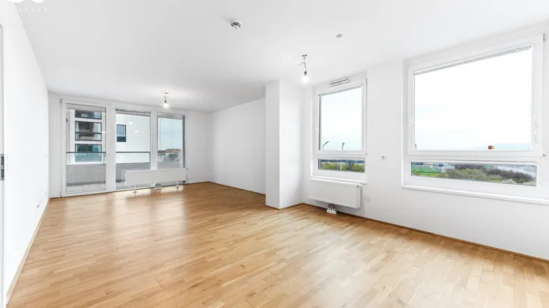Helles, leeres Wohnzimmer mit Parkettboden, großen Fenstern und Zugang zum Balkon.