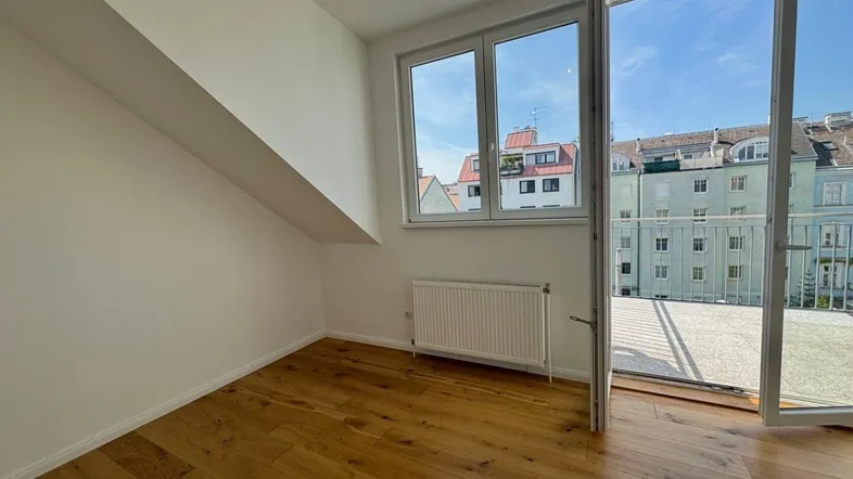 Helles Zimmer im Dachgeschoss mit Holzboden, schräger Decke und Zugang zu einem Balkon.