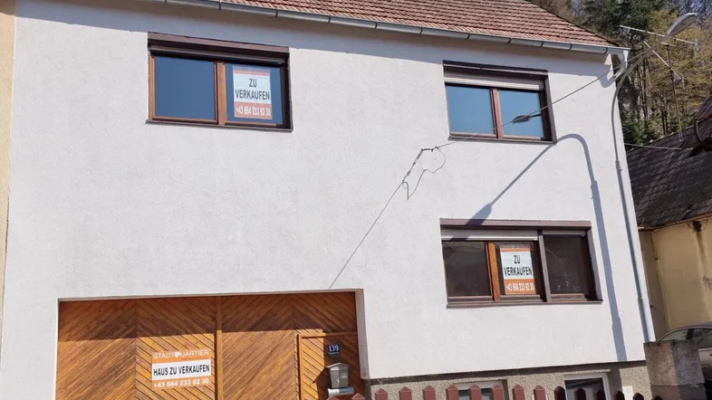 Weiß verputztes Einfamilienhaus mit braunen Fenstern und "Zu verkaufen"-Schildern.