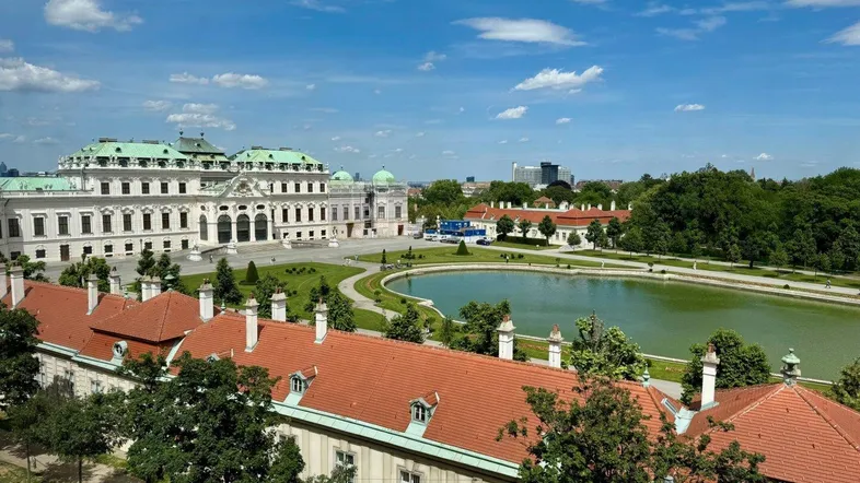 Panoramablick auf ein historisches Schloss mit weitläufigen Gärten und einem Teich unter blauem Himmel.