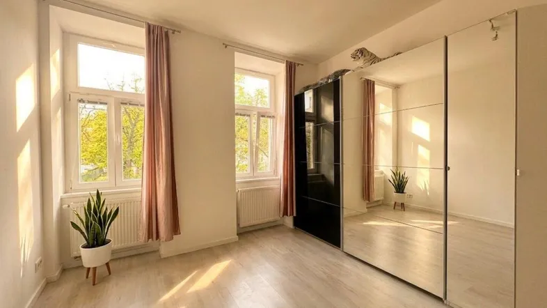 Helles Zimmer mit großen Fenstern, hellem Boden und einem geräumigen Spiegelschrank, durchflutet von Sonnenlicht.