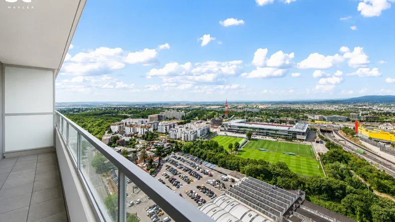 Geräumiger Balkon mit Glasgeländer und weitem Blick über die Stadt und Sportanlagen.