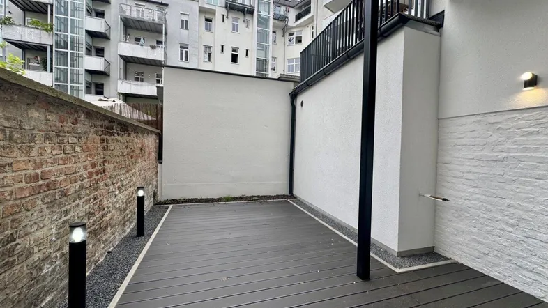 Moderne Terrasse mit dunklem Holzdeck, umgeben von einer Ziegelmauer und weißen Wänden in urbaner Umgebung.