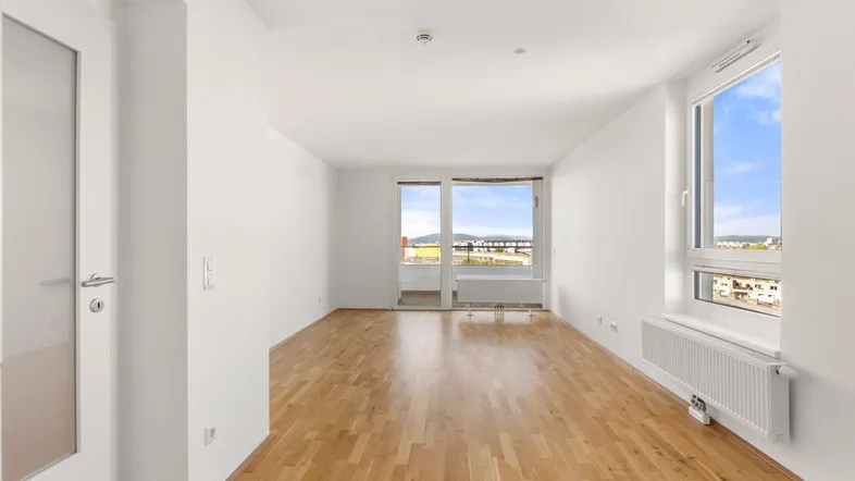 Helles, leeres Wohnzimmer mit Parkettboden und Zugang zu einem Balkon mit Weitblick.