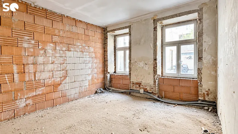 Innenraum im Rohbauzustand mit unverputzten Ziegelwänden und zwei Fenstern, bereit für die Renovierung.