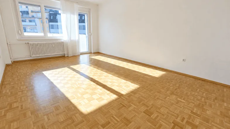 Helles, leeres Zimmer mit Parkettboden und Zugang zu einem Balkon, durchflutet von Sonnenlicht.