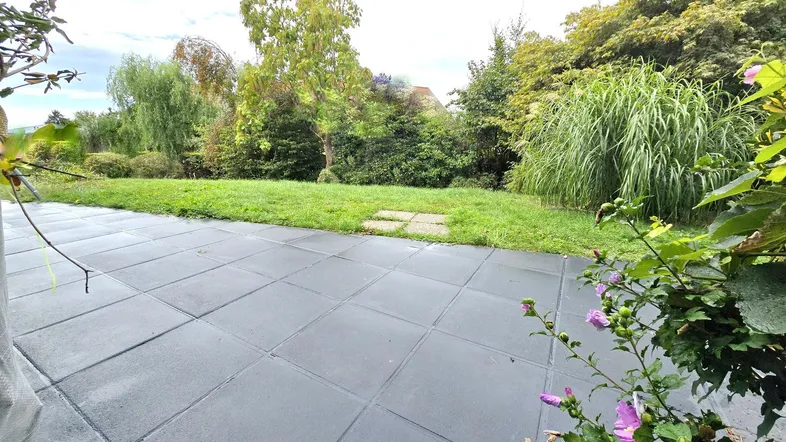 Große gepflasterte Terrasse mit angrenzendem Rasen und üppiger grüner Bepflanzung im Hintergrund.