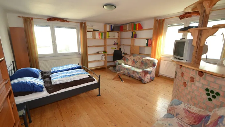 Geräumiges Schlafzimmer mit Holzdielenboden, zwei Fenstern, Sofa und Regalen.