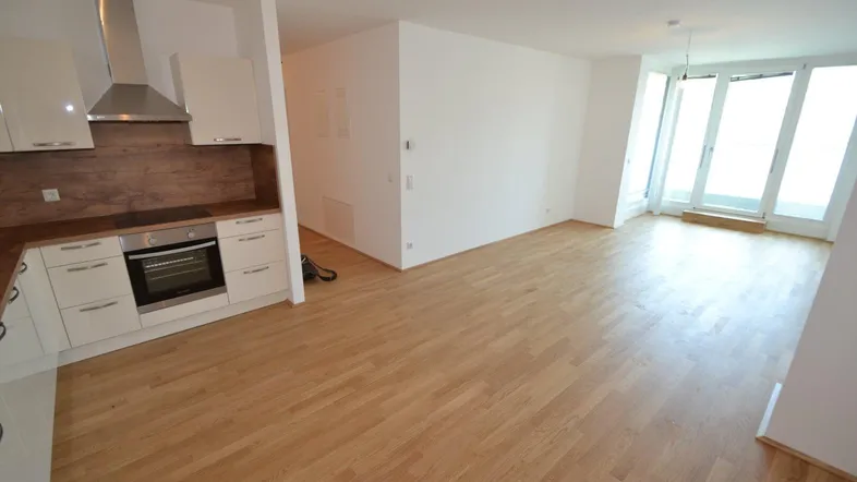 Heller offener Wohnbereich mit integrierter Küche, Parkettboden und Zugang zum Balkon.