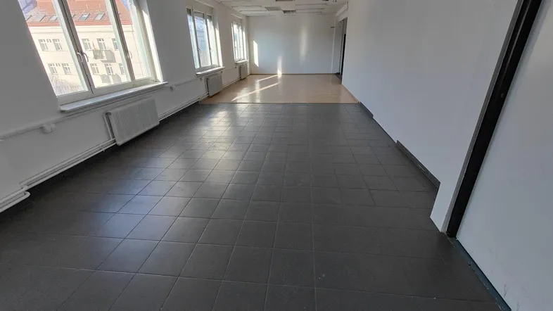 Geräumiges, leeres Büro mit großen Fenstern, Fliesenboden und viel Tageslicht.