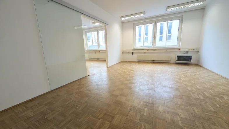 Geräumiges Büro mit Fischgrätparkett und großen Fenstern, durch eine Glasschiebetür abgetrennt.
