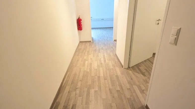 Langer Flur mit hellem Holzoptikboden, weißen Wänden und offener Tür zu weiteren Räumen.