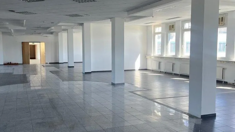 Großer, heller Büroraum mit Säulen, Fliesenboden und vielen Fenstern, die Tageslicht hereinlassen.